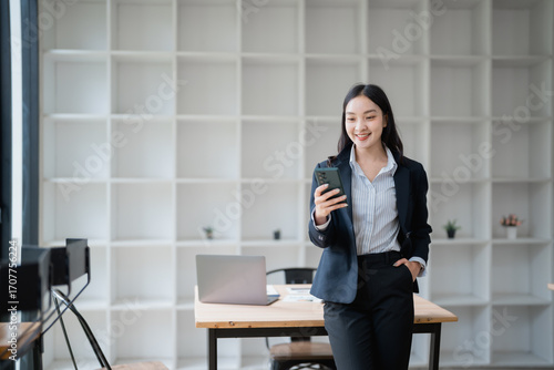 Young Asian businesswoman smiling while using her smartphone, leaning against a desk in a modern office filled with natural light, surrounded by a laptop and important documents Free Download Young Asian businesswoman smiling while using her smartphone, leaning against a desk in a modern office filled with natural light, surrounded by a laptop and important documents - GFXLoad