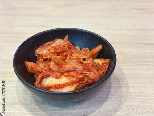 Authentic Korean Horizontal Shot: Spicy Fermented Napa Cabbage Kimchi (Baechu Kimchi) Served as a Side Dish in a Matte Black Bowl on a Light Wooden Table.