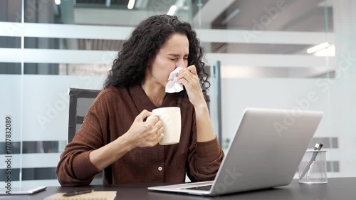 Sick young businesswoman suffers from a cold sitting at a workplace in a business office. Female employee coughs, sneezes, blows her nose with a handkerchief, drinks a hot drink. The woman has the flu