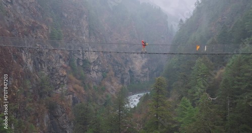 Drone view of Hillary Bridge near Monjo on Everest Base Camp trek, Nepal. A tall suspension bridge in rain shows tourists crossing, looking down at river and Himalayan landscape with awe and thrill