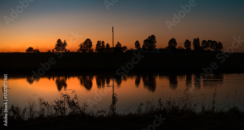sunset over the horizon with the reflection of trees and a tower in the calm ...