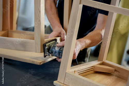 A craftsman sands the wooden frame of a chair with an orbital sander during the production process. The image shows woodworking and artisanal furniture.