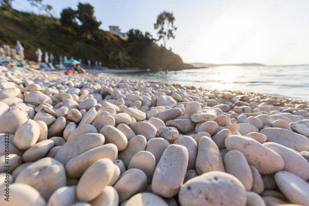 Obraz premium An up-close, low-angle shot of smooth, sun-warmed white pebbles on a beach with the bright sun creating a beautiful lens flare over the gently moving, blurred ocean.