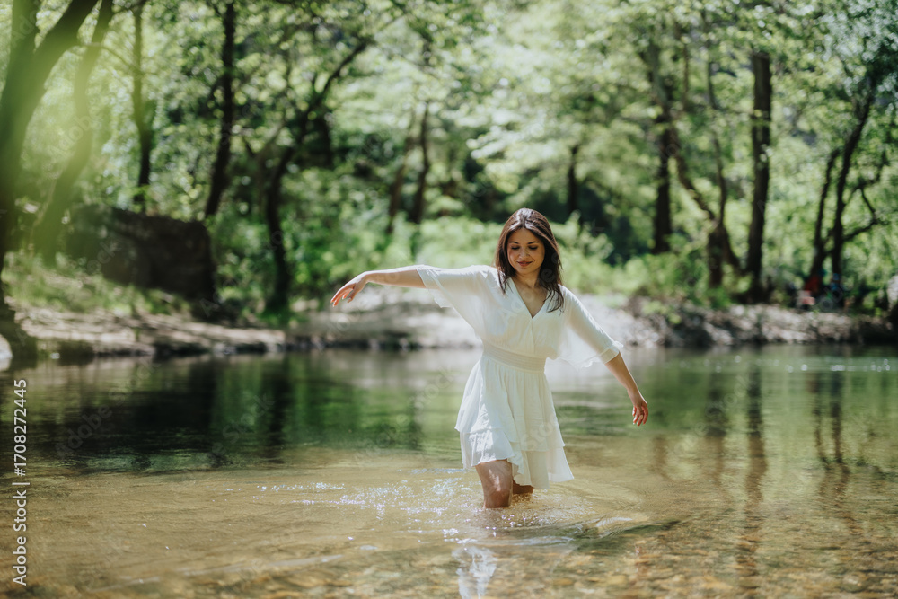 Fototapeta premium A woman dressed in white wading in a clear forest stream, surrounded by lush greenery. The atmosphere conveys peace and connection with the outdoors, making for a refreshing and vibrant scene.