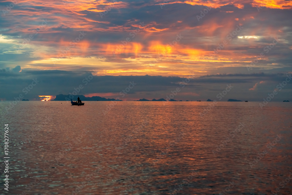 Fototapeta premium Small Boat Floating on Calm Sea During Dramatic Sunset with Fiery Sky and Island Silhouettes on Horizon, captured in wide angle, Concept Tranquil Evening Seascape