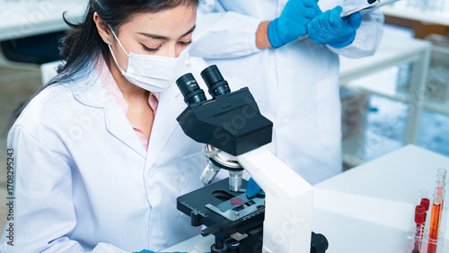 Female scientist examining a blood sample through a microscope while a colleague records notes, representing healthcare diagnostics, virus testing, laboratory research, and scientific innovation.