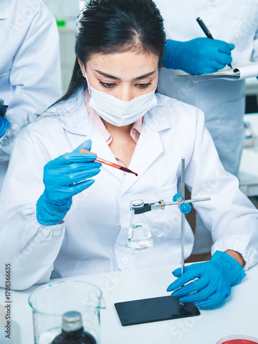 A female scientist carefully drops liquid into a flask while colleagues take notes and observe through a microscope, symbolizing teamwork, research, and the pursuit of answers in science and medicine.
