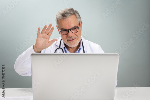 A doctor in a white coat engages in an online consultation. He smiles and waves at the camera, creating a friendly atmosphere for the virtual visit.
