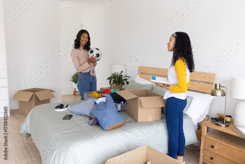 Diverse mom and teenage daughter unpacking moving boxes around bed in bedroom holding soccer ball