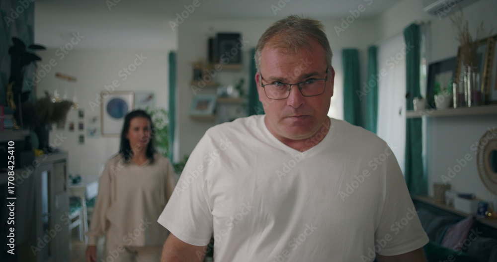 Fototapeta premium Man staring at camera with intense expression during argument, woman in background in living room, tense and emotional moment of relationship conflict and frustration