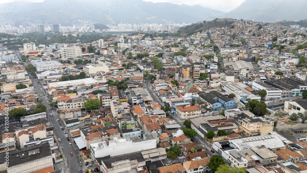 Fototapeta premium Imagem aérea de área residencial próxima ao Estádio São Januário, no Rio de Janeiro, com ruas, casas, galpões, prédios e morros ao fundo sob céu cinzento e atmosfera urbana.
