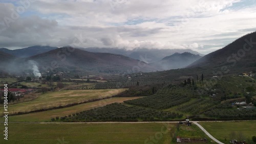 Aerial view shot of Italian countryside during the cloudy day, mountain peaks covered with light mist, rising smoke from burning dry leaves, rustic fields for cultivation, plowing and food production