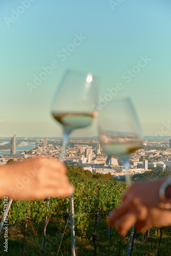 Vertical photo of vineyard rows and Vienna skyline under warm sunlight with bright blue sky. Foreground shows blurred male and female hands clinking wine glasses, creating romantic, cozy atmosphere.