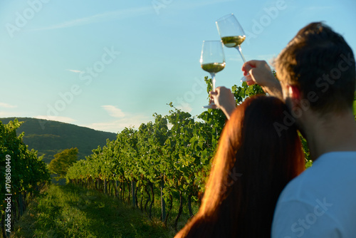 Horizontal photo of a couple from behind, heads close together, holding white wine glasses above vineyard rows. Warm golden sunset light creates romantic, cozy, and intimate outdoor wine atmosphere.