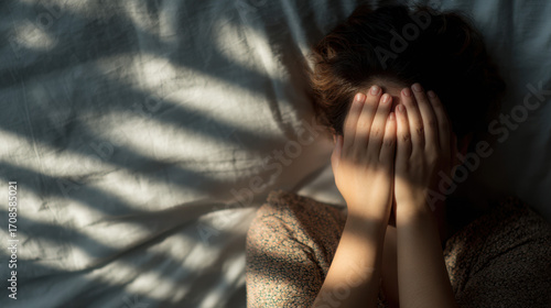 Young woman covering her face while lying in bed, expressing sadness or emotional distress through body language and shadow play.
