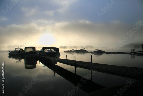 Boats in the morning mist along the pier. Sunrise behind the clouds.