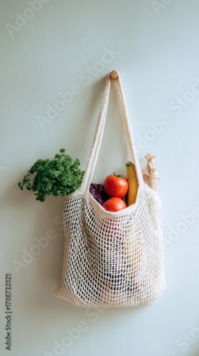 Reusable cotton mesh bag filled with fresh produce including tomatoes, bananas, and parsley hanging on white wall, zero waste lifestyle concept.
