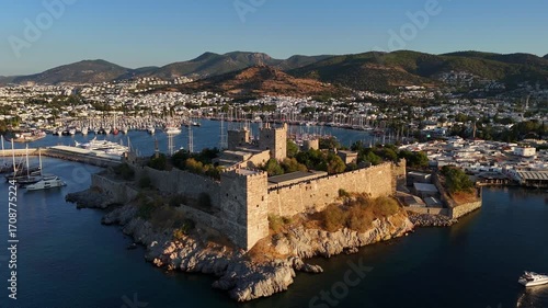 Drone aerial view of Bodrum Castle on the Aegean coast of Turkey at sunrise, with a marina full of yachts, white houses, and green hills in the background.