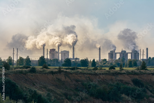 Dolomite processing plant in Belarus with multiple smokestacks emitting thick white smoke, surrounded by green fields and trees under a hazy sky.