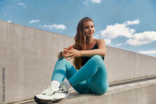 Fit young woman in athletic wear sits on a concrete ledge, showcasing a relaxed workout pose against a bright blue sky, embodying a healthy lifestyle and fitness motivation.