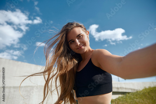 Fit young woman with long hair takes a selfie outdoors, showcasing her athletic physique in a black sports top against a bright blue sky with fluffy clouds, capturing a moment of joy.