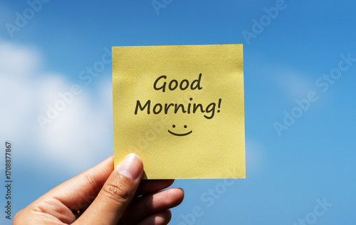 Schilderij op canvas Hand holding yellow sticky note with text Good Morning! and smiley face against clear blue sky background