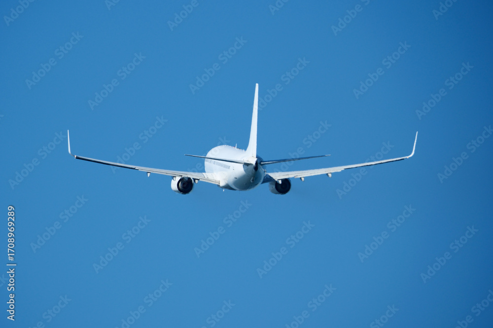 Fototapeta premium The plane that's just taken off drifts away from the airport across clear blue sky. Airplane flying in the blue sky on a sunny day. Airplane taking off, rear view. Travel concept.