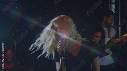 Lead singer stepping forward holding mic singing while band strumming guitar under light bursts