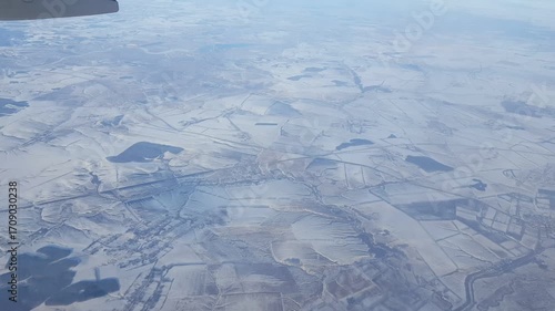 Snowy land below camera tilts up passenger window view from plane.