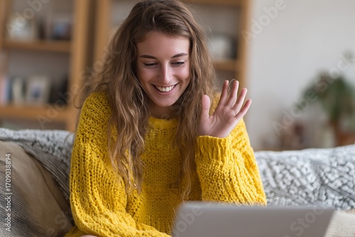 Cheerful Young Woman In A Cozy Yellow Sweater Smiles And Waves While Having A Video Call On Her Laptop From Her Sofa At Home, Virtual Communication, Remote Connection