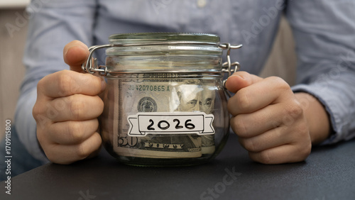 Unrecognizable woman holding Saving Money In Glass Jar filled with Dollars banknotes. 2026 year transcription in front of jar. Managing personal finances extra income for future insecurity background