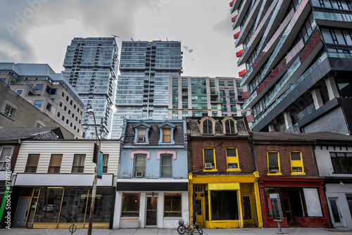 Row of older storefront houses on Queen Street in Toronto Canada set against modern condo towers, showing the contrast between heritage streetscape and high-rise redevelopment in the downtown core.