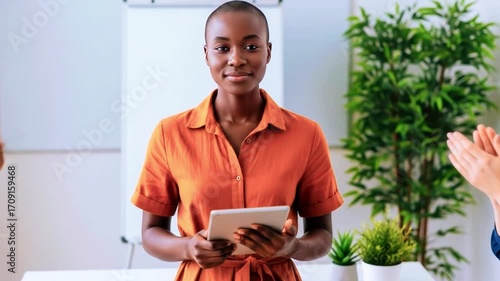 Confident woman in an orange dress holding a tablet, receiving applause in a bright office setting