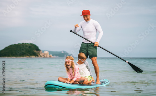 Dad paddles while kids sit in front of him on stand-up paddleboard, having fun on calm ocean waves. Family activity, connection, and laughter in motion.