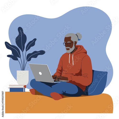 A stylish senior Black man with a beard and glasses sits comfortably cross-legged, focused on his laptop, working remotely from home in a modern, cozy setting.