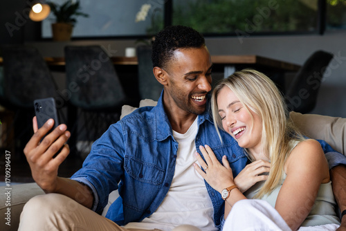 Fotografi Couple sitting on couch laughing while taking selfie with smartphone at home