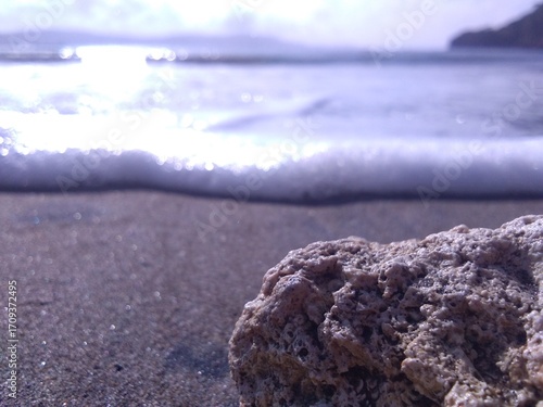 A close-up of rugged rocks in the foreground with foamy waves rolling onto a dark, sandy beach under a bright blue sky