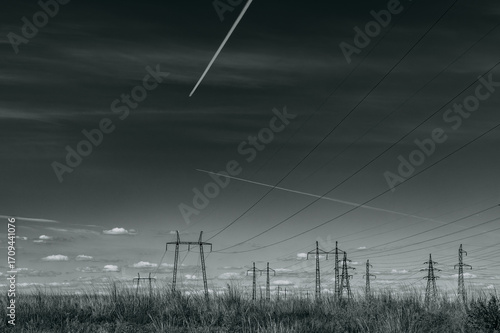Black and white landscape. Above the wires of a high-voltage power line, the sky is filled with white trails from high-flying airplanes