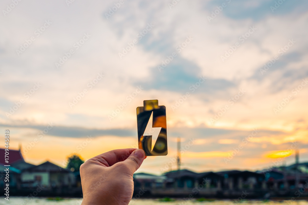 Fototapeta premium Hand holding battery icon with lightning bolt in front of river village at sunset, symbolizing clean energy, power innovation, and sustainable development. Ideal for eco-tech and green power concepts.