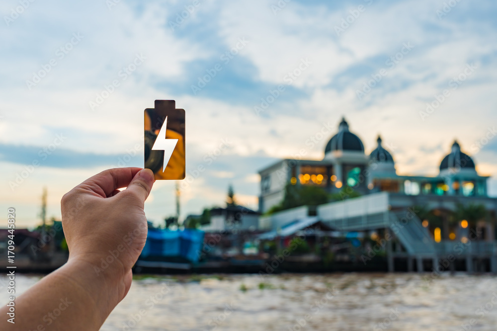 Fototapeta premium Hand holding golden battery icon with lightning bolt against cityscape background, symbolizing energy, electricity, and charging. Perfect for sustainable power, smart city, or eco technology concept.