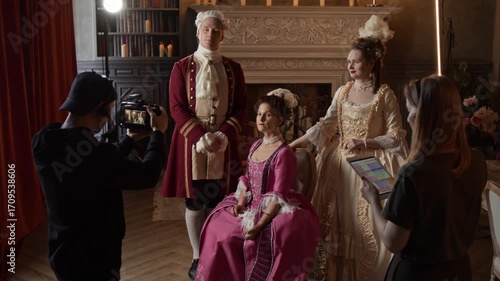 Medium full shot of male videographer shooting portrait of noble aristocratic family on set of historical movie, actors in 18th century costumes posing, and female producer holding tablet with script