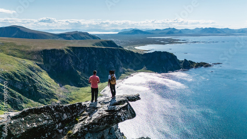 Tableau sur toile Two hikers stand on a rocky outcrop at Matind Mountain, overlooking the beautiful coastline of Bleik, Andoya