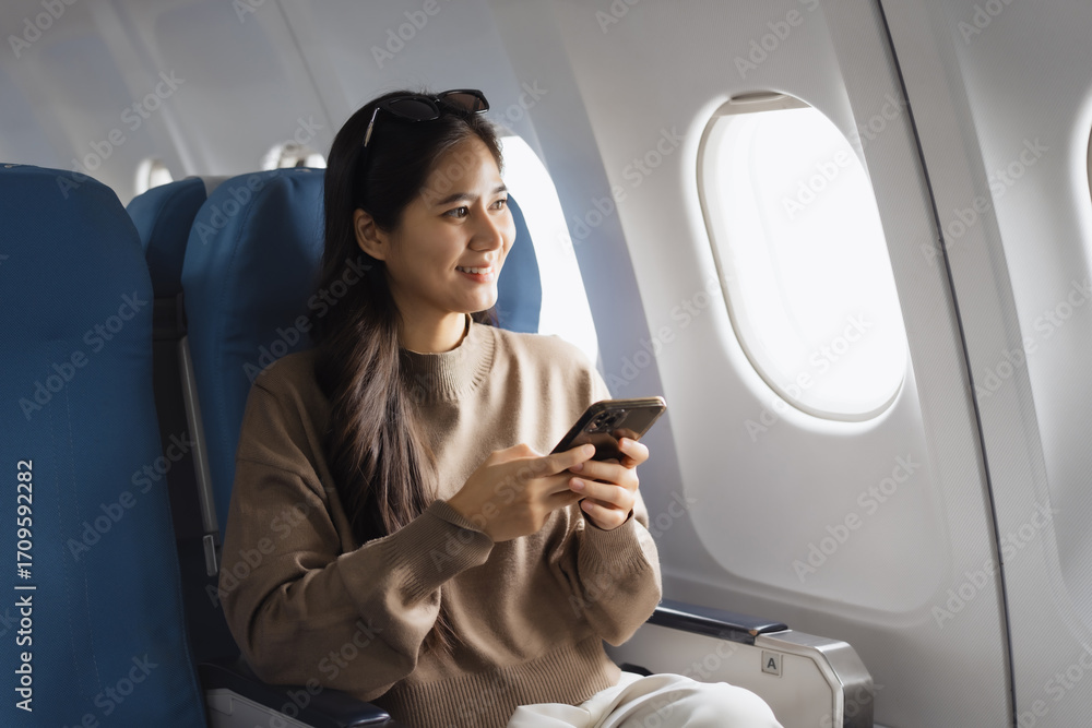 Fototapeta premium An Asian woman sits comfortably on an airplane, holding her smartphone smiling while looking out the window, representing modern travel,connectivity, leisure,the joy of experiencing a flight journey