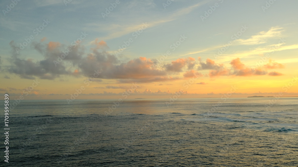 Obraz premium A peaceful scene of the calm sea stretching to the horizon under a sky with soft, pastel-colored clouds. A small island is visible in the distance. The image conveys a sense of serenity