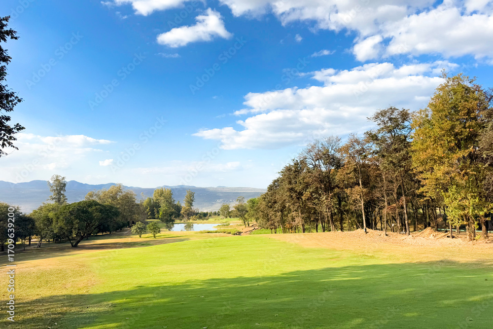 Naklejka premium Beautiful golf course view with mountains, forest, and bright sky, showing a scenic fairway and sand bunker.