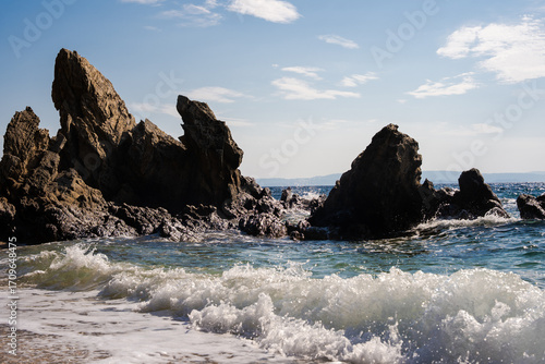 Dramatic seascape with jagged rocks rising from the clear blue water, waves crashing near a small sandy beach on the vibrant Greek coast.