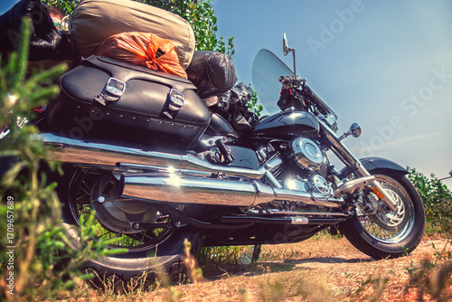 Schilderij op canvas A black cruiser motorcycle, heavily loaded with travel gear, parked on a dirt road under a clear blue sky