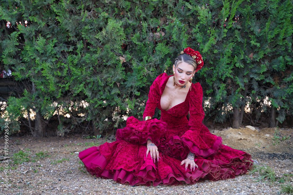 Fototapeta premium Young, beautiful, blonde woman in red flamenco dress, flowers in her hair and gold jewelry, performing flamenco poses sitting on the ground with green plants in the background. Concept dance, art.