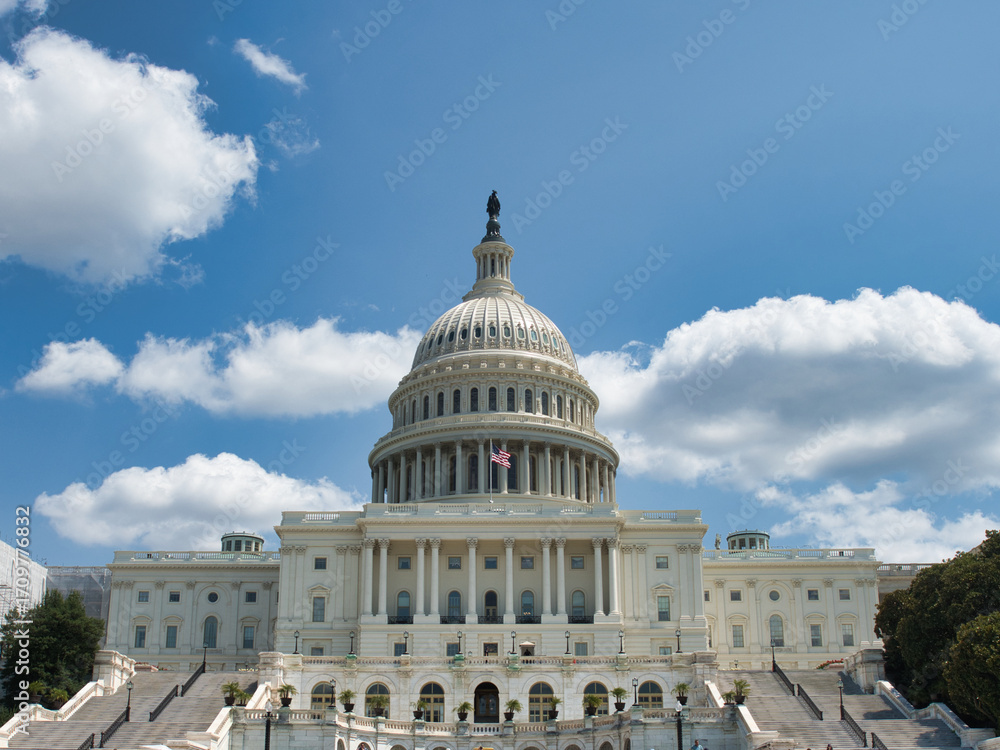 Naklejka premium United States Capitol Building Main Facade - The historic U.S. Capitol Building with its grand staircase and dome against a clear blue sky with white clouds.