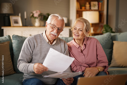 Photos An elderly man wearing glasses reads paperwork while sitting with his wife on a sofa